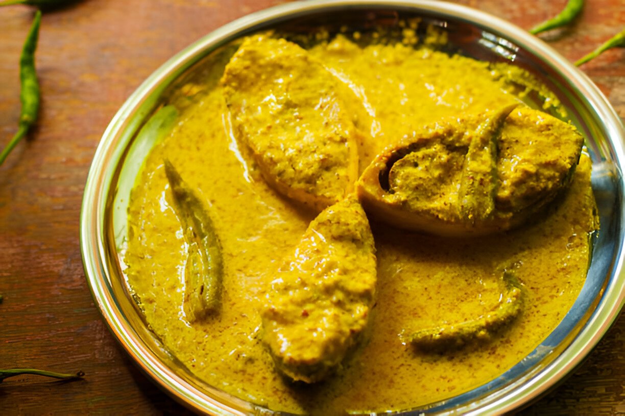 Close-up of a fish curry in a yellow mustard gravy, served in a steel plate.